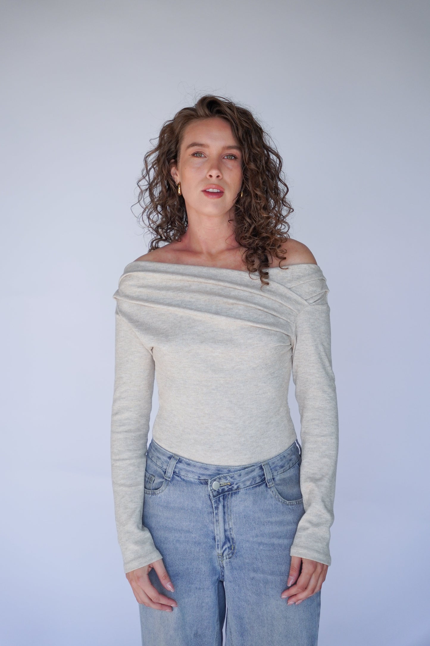 Woman wearing a light gray off-shoulder sweater and blue jeans against a plain background.
