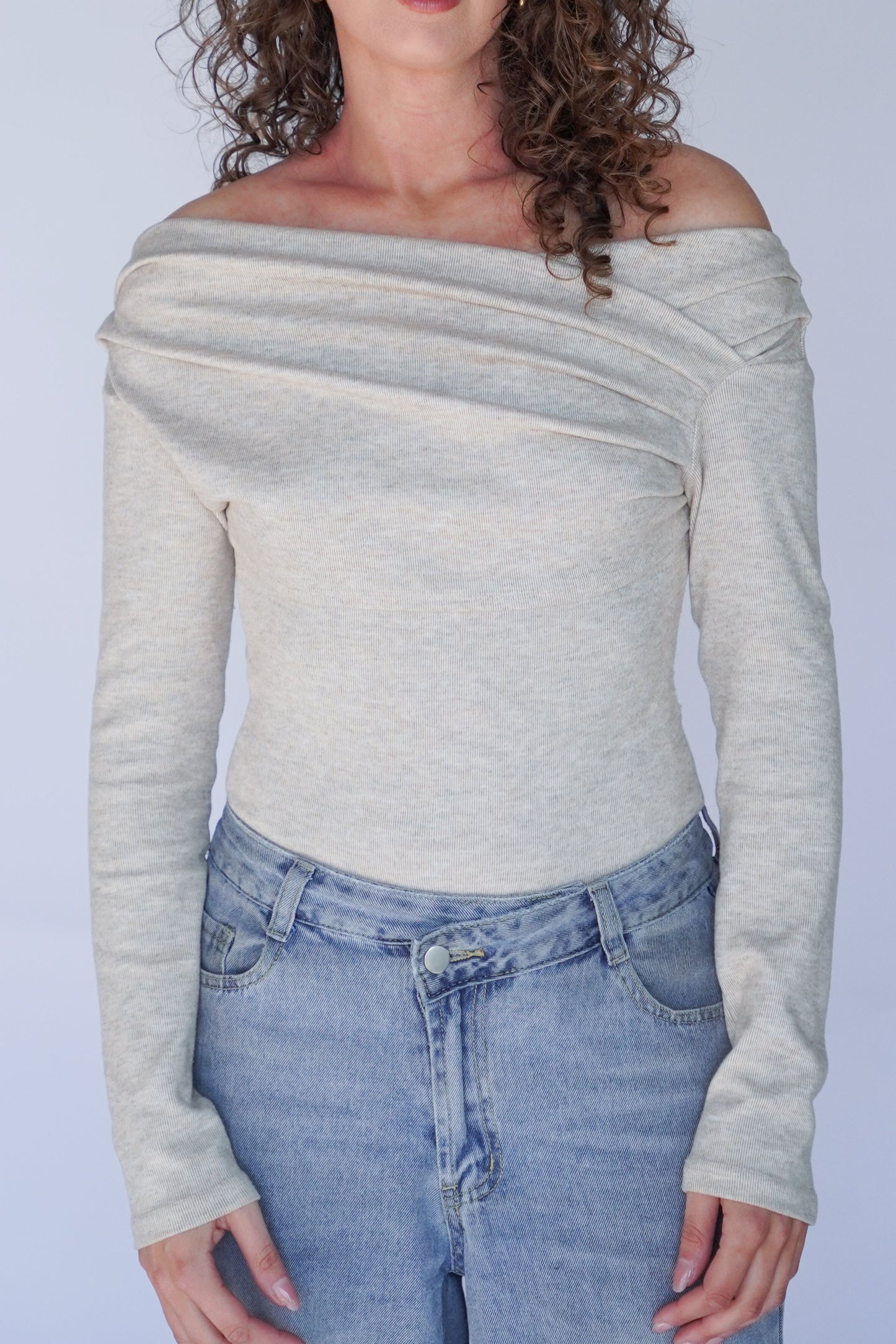 Woman wearing a beige off-shoulder top and blue jeans against a plain background.