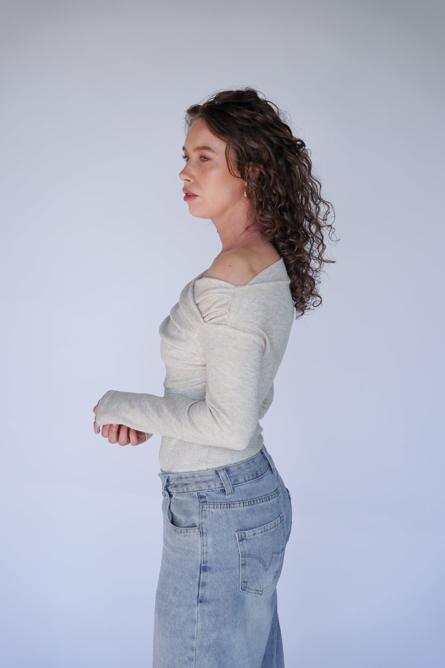 Person wearing a light beige off-shoulder top and blue jeans against a plain background.