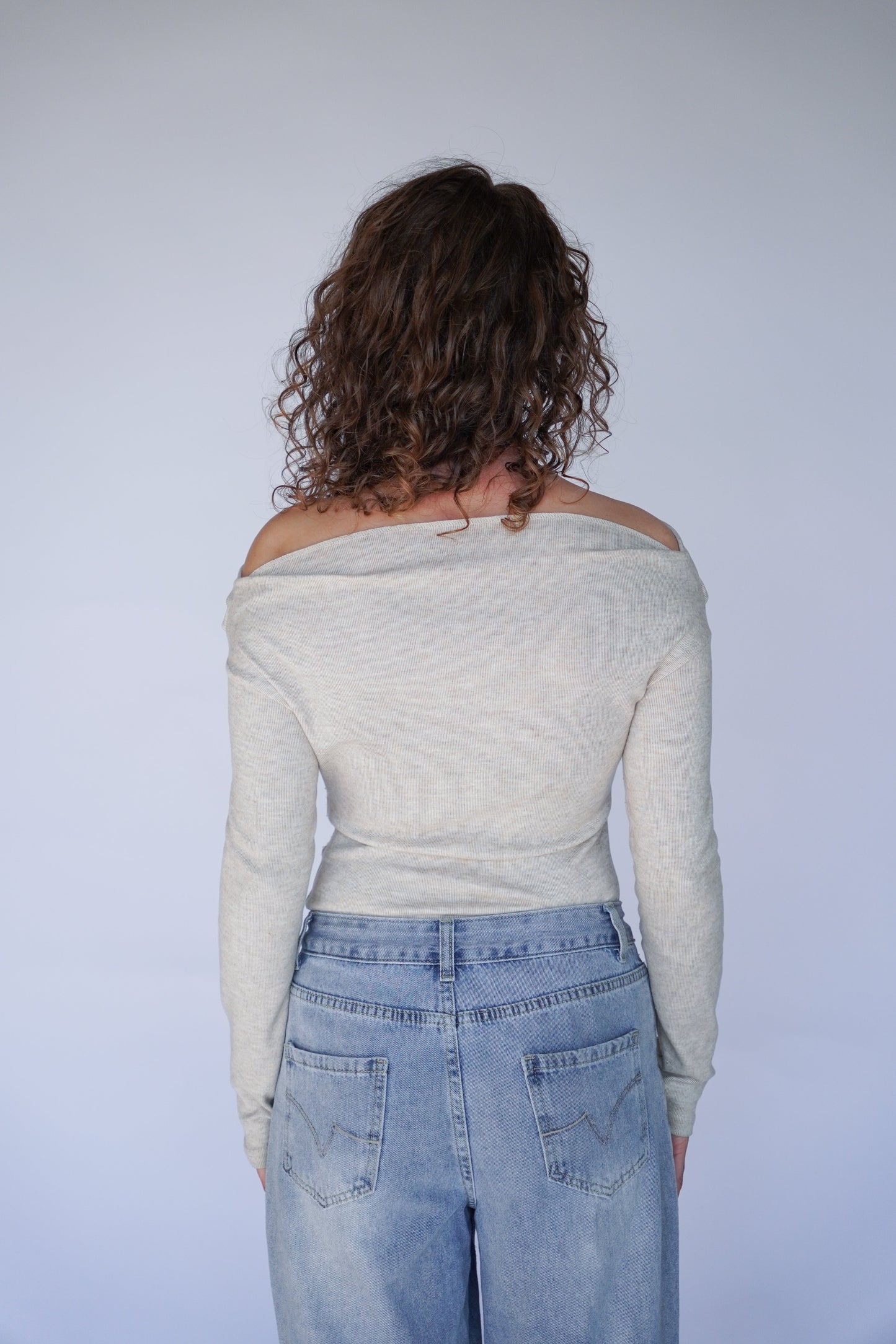 Person wearing a light beige off-shoulder top and blue jeans against a plain background.