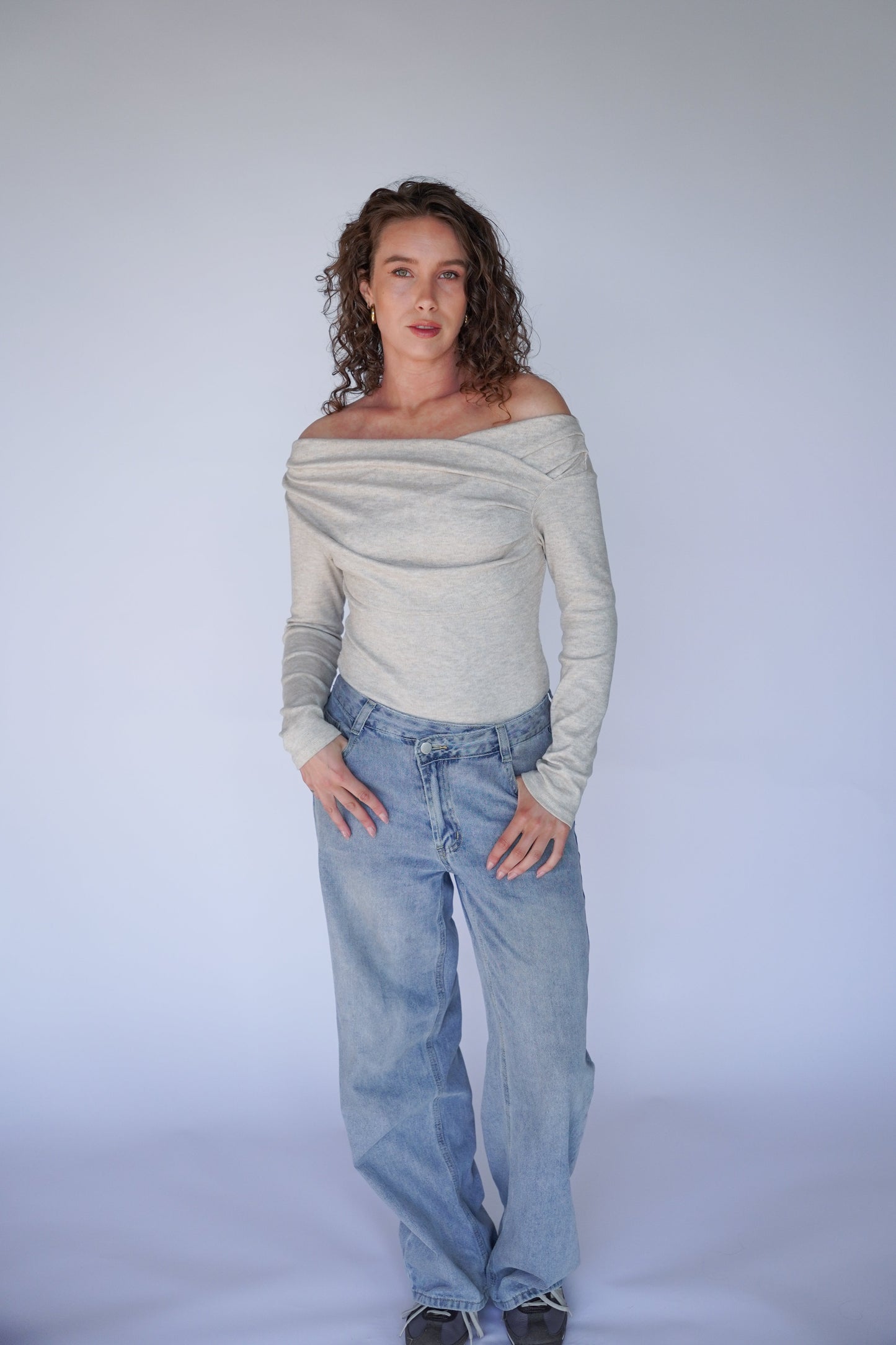Woman wearing a gray off-shoulder top and blue jeans against a plain background.