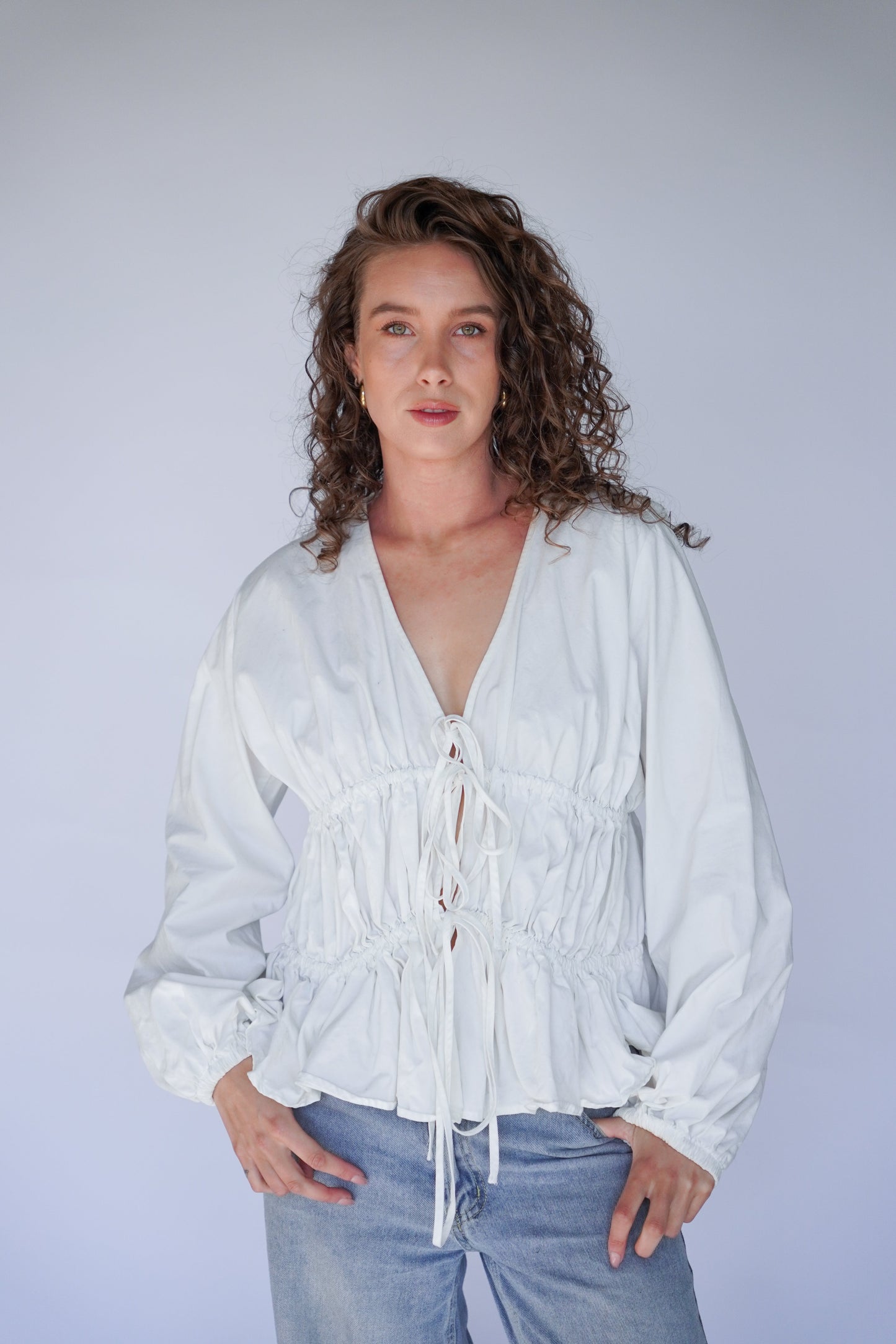 Woman wearing a white blouse with lace-up front and blue jeans against a plain background.