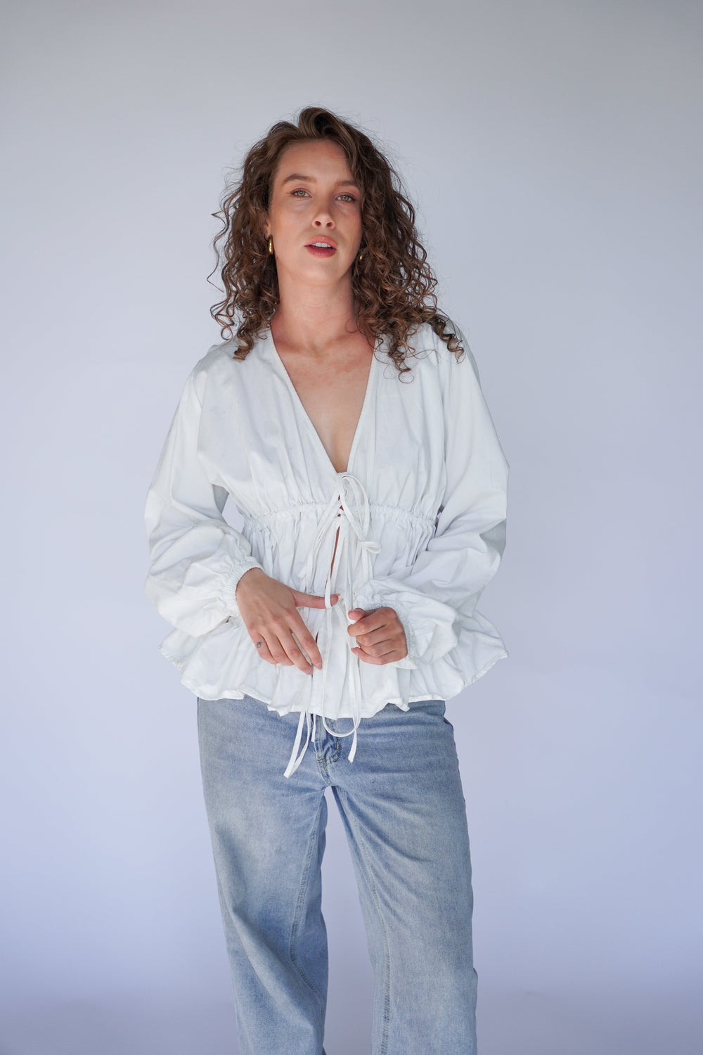 A woman standing, wearing a white Andie blouse with balloon sleeves and tie-front details, paired with blue jeans.