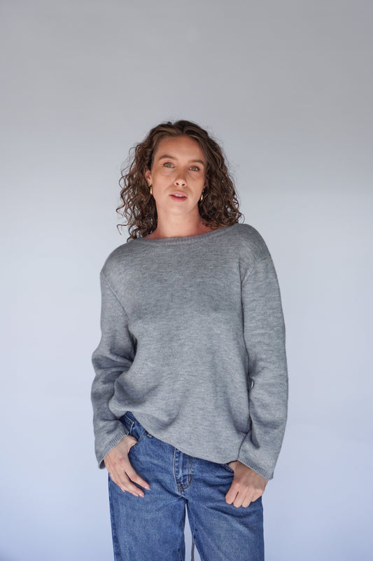 A female model wearing a gray Bella knit sweater with long sleeves and standing against a plain background.