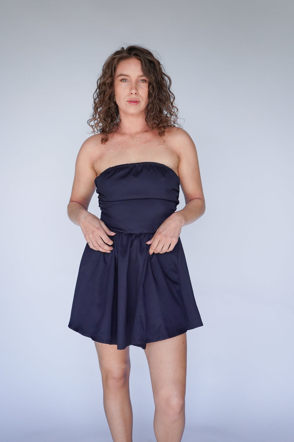 A woman standing, wearing a strapless, navy-colored mini dress with a tie detail at the back and a tiered skirt.