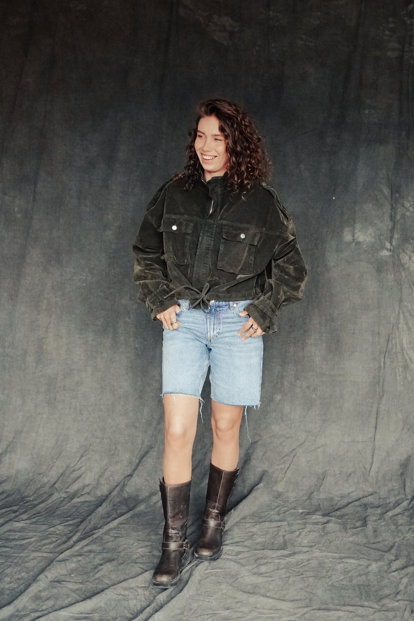 Person wearing a dark jacket, light blue shorts, and boots against a gray background.