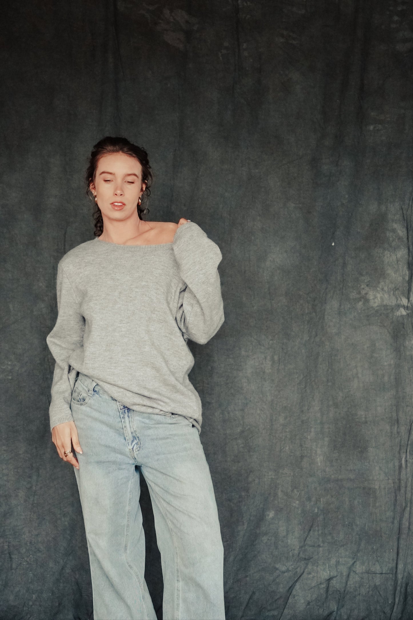 Person wearing a gray sweater and light blue jeans against a dark background.