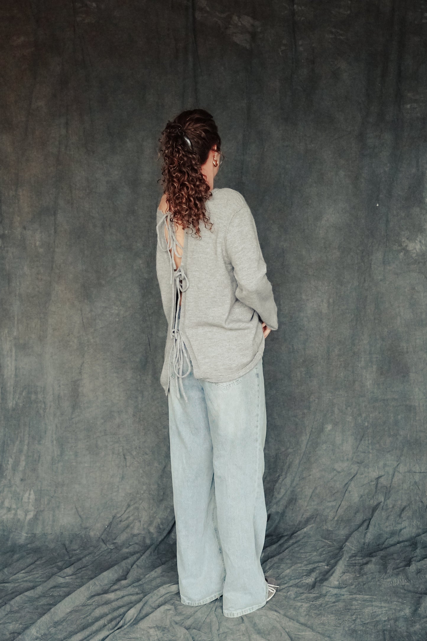 Person wearing a gray cardigan and light-colored pants against a textured gray background.