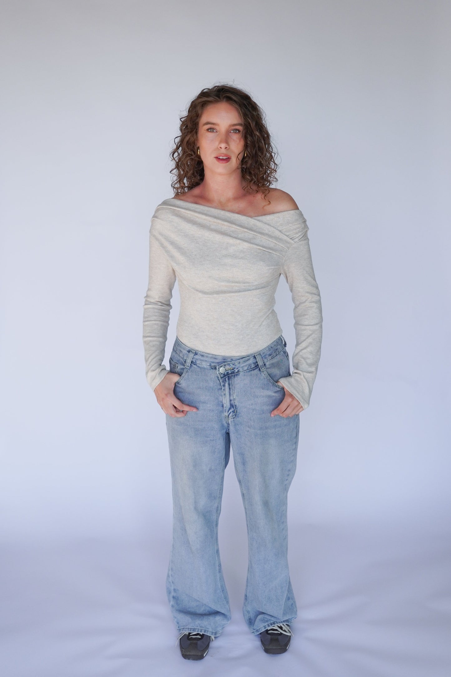 Woman wearing a gray off-shoulder top and blue jeans against a white background