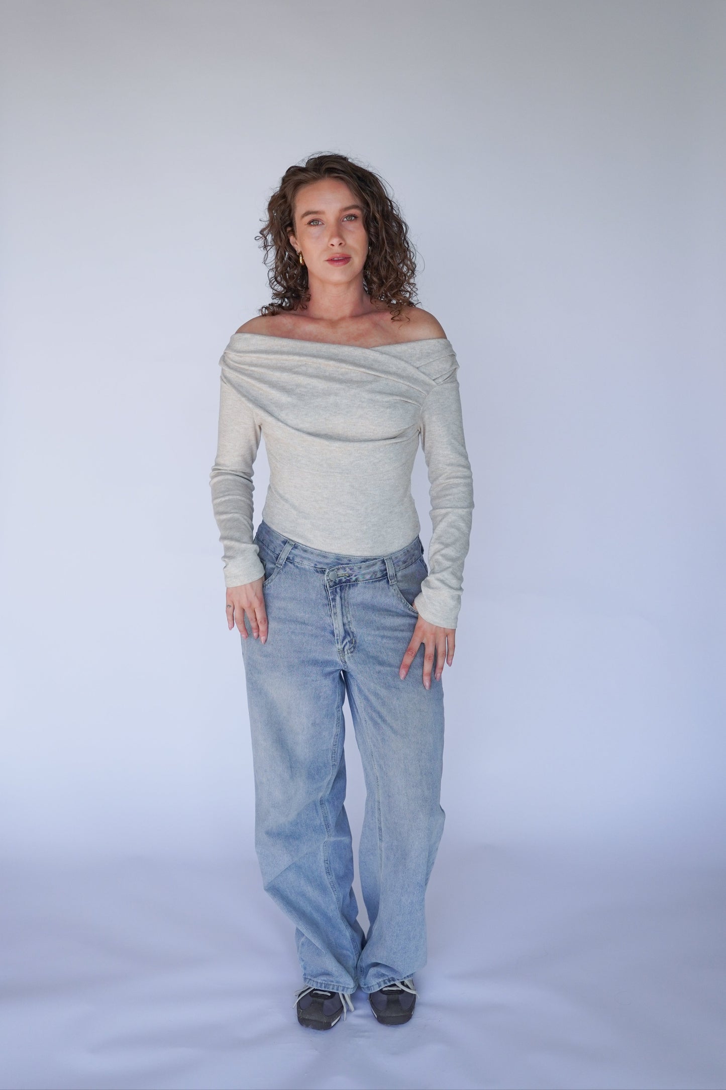 Woman wearing a light gray off-shoulder top and blue jeans against a plain background.