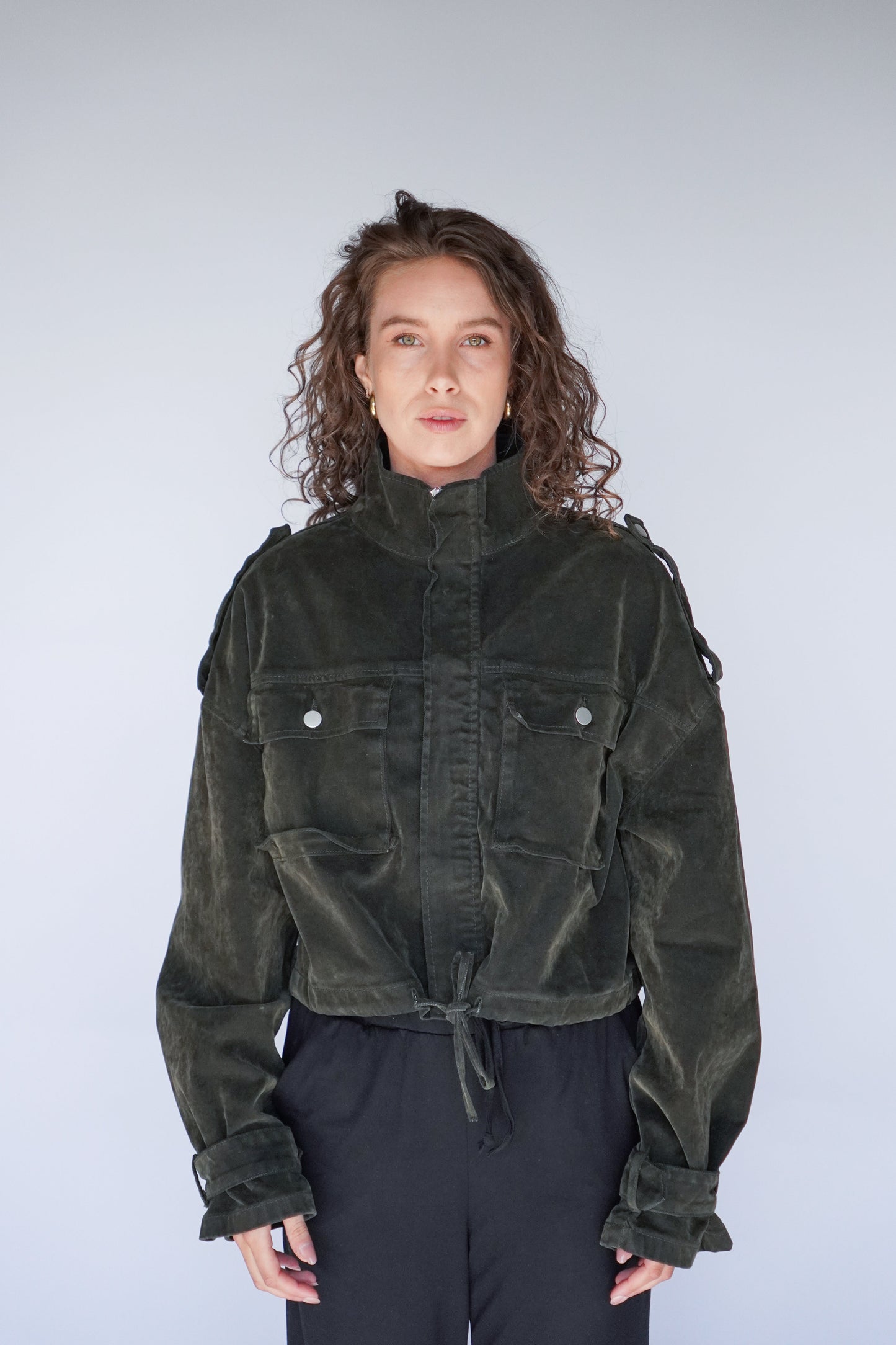 A woman is standing, wearing a green cropped jacket with a suede-look finish over a black top and black pants.