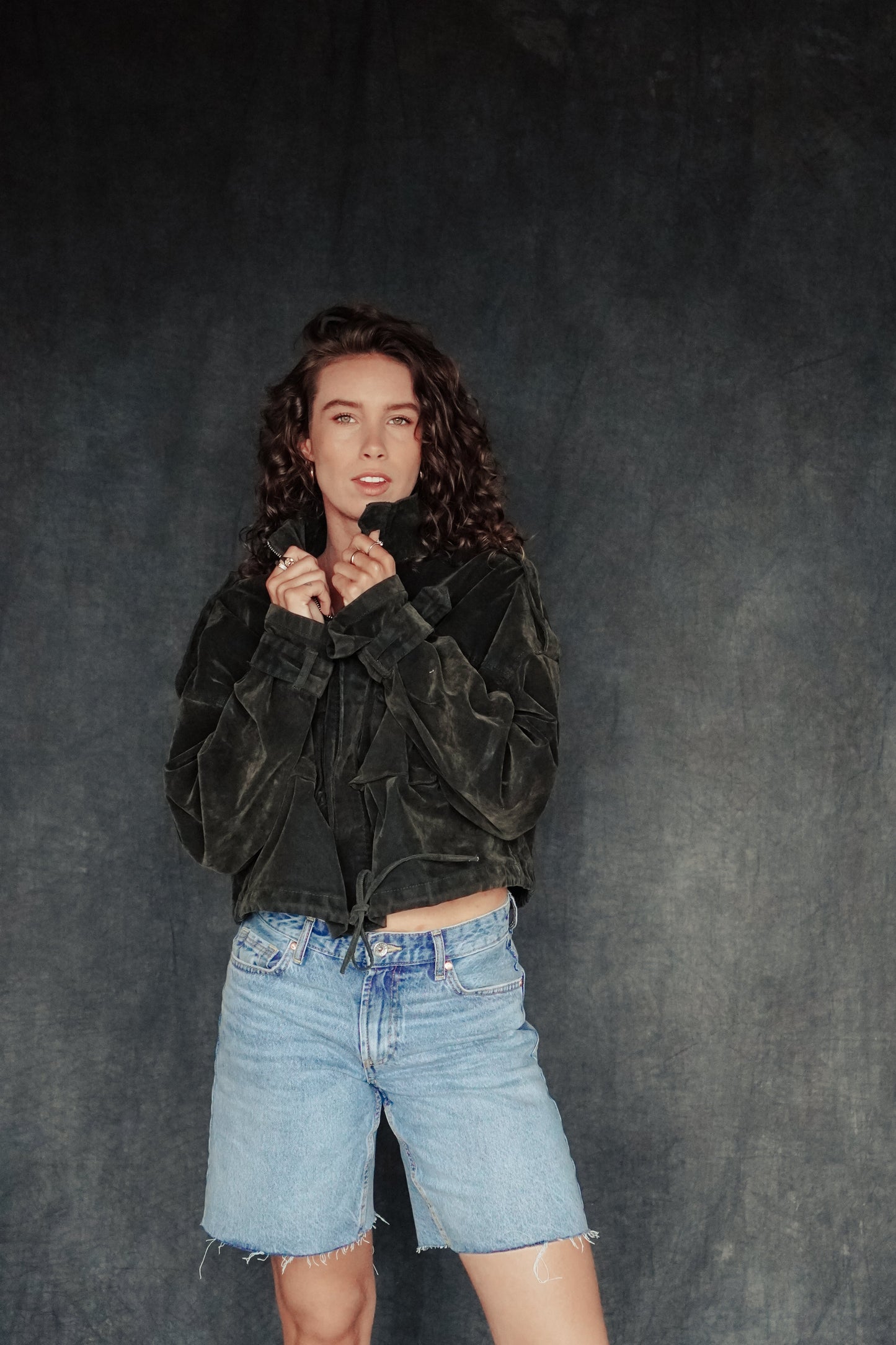 Person wearing a green suede jacket and denim shorts against a dark background