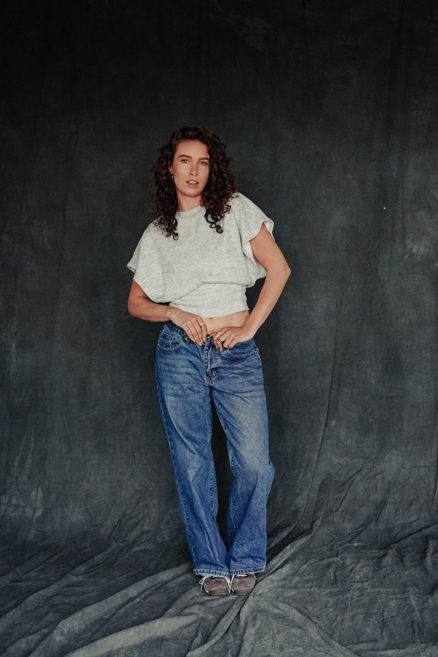 A person wearing a white cropped top and blue jeans standing against a grey background.