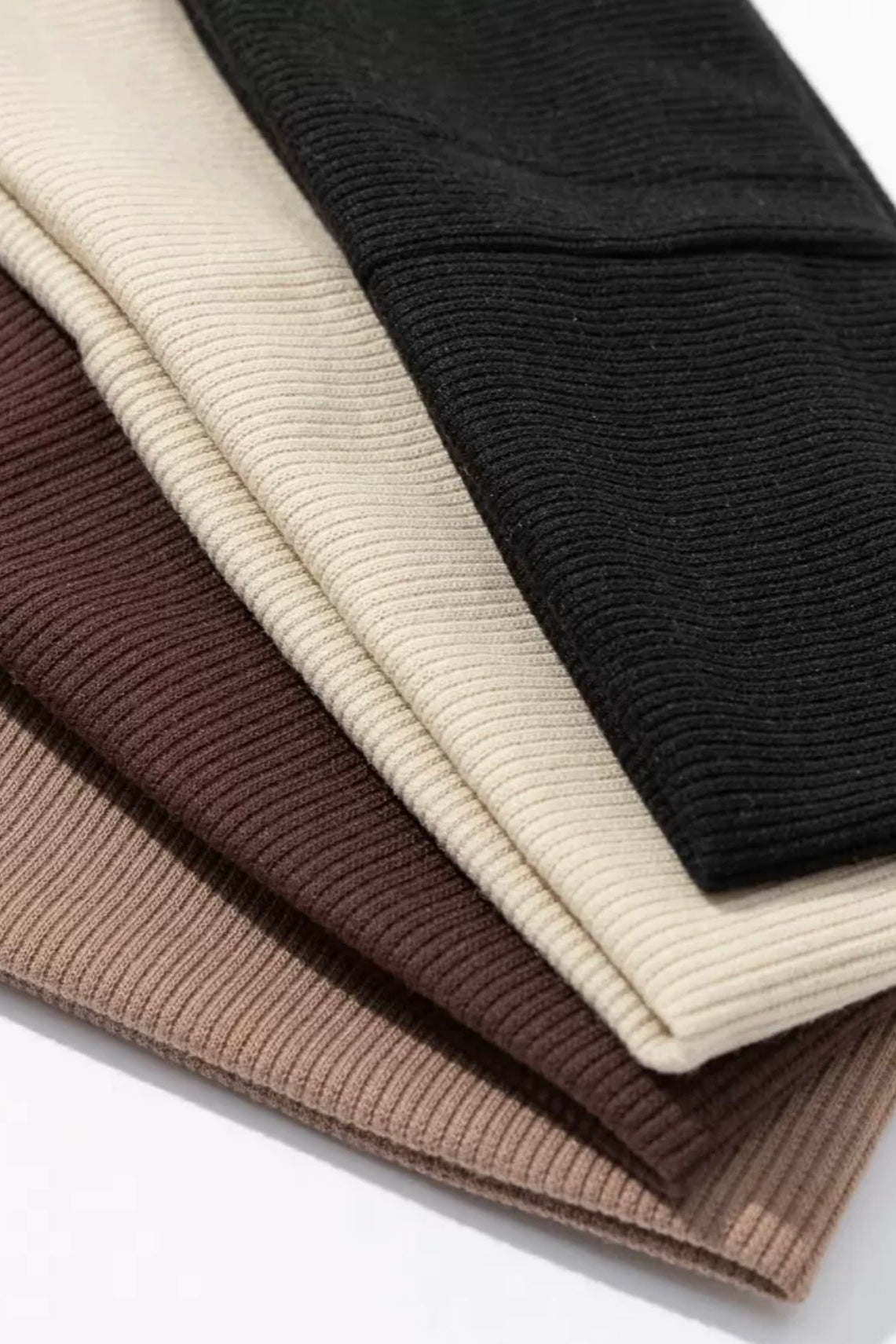 Stack of folded ribbed knit sweaters in black, beige, and brown on a white background