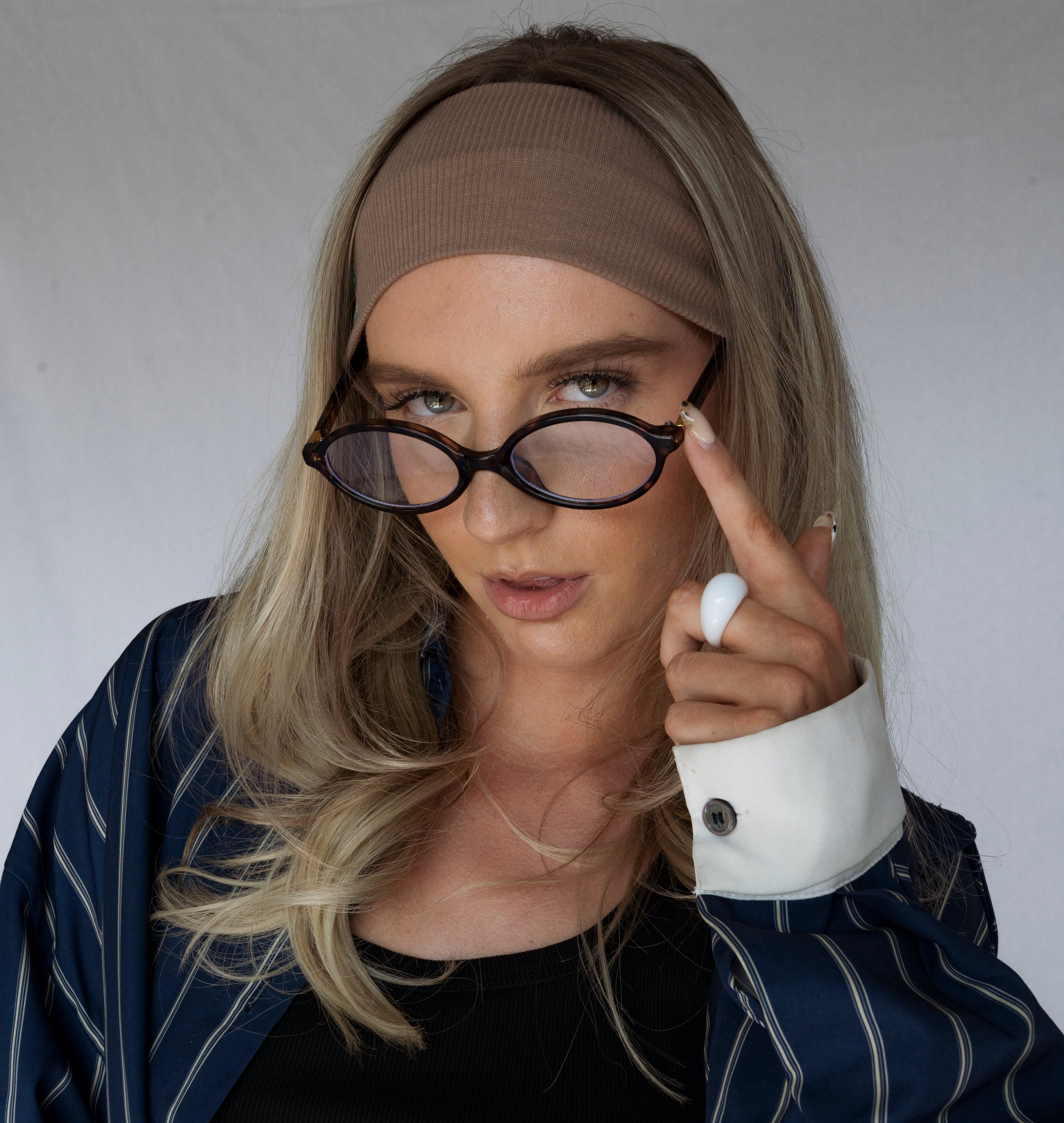 Person wearing a brown headband and pointing to their eyeglasses against a plain background